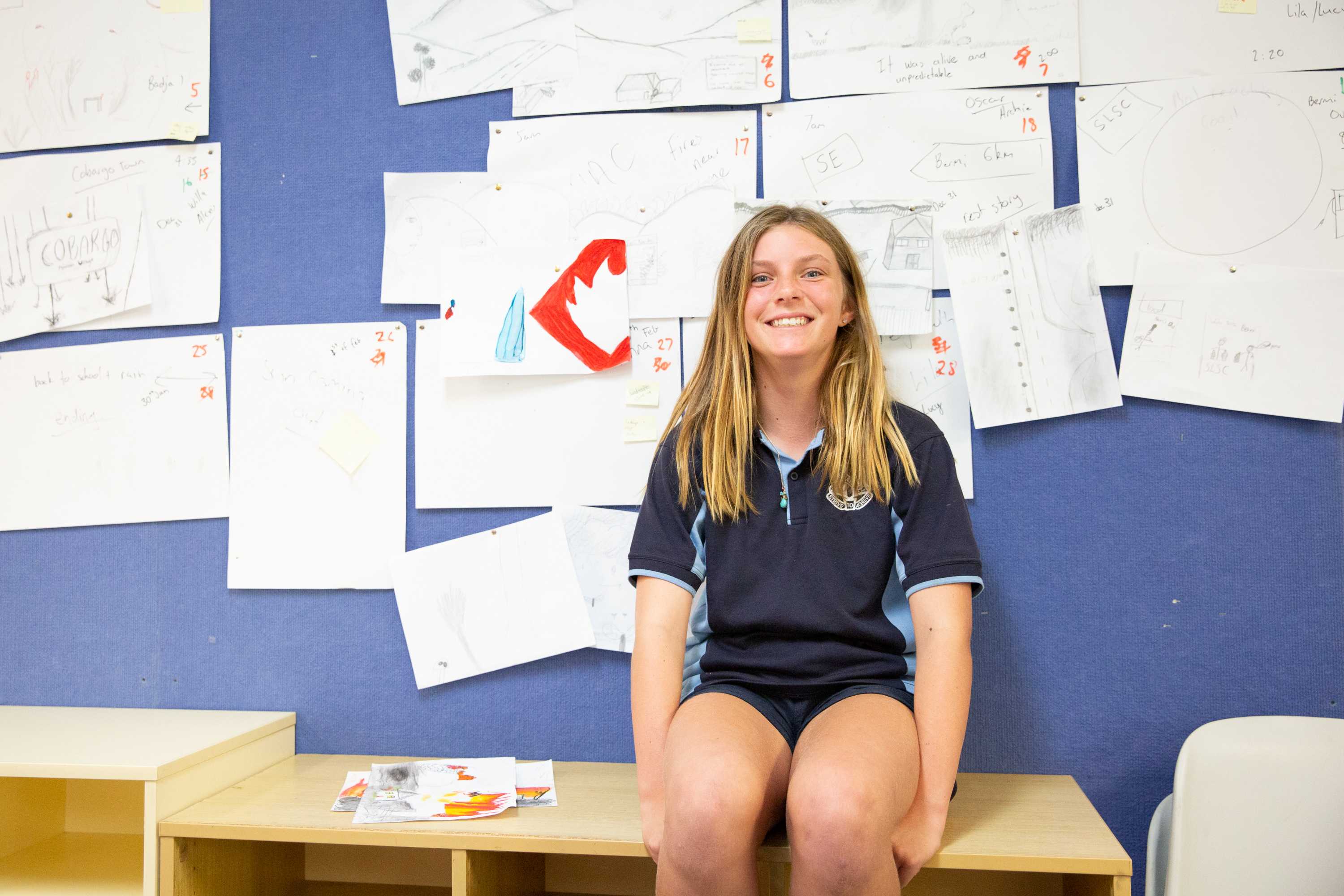 A girl in a school uniform sitting in front of a blue wall with children's art on it