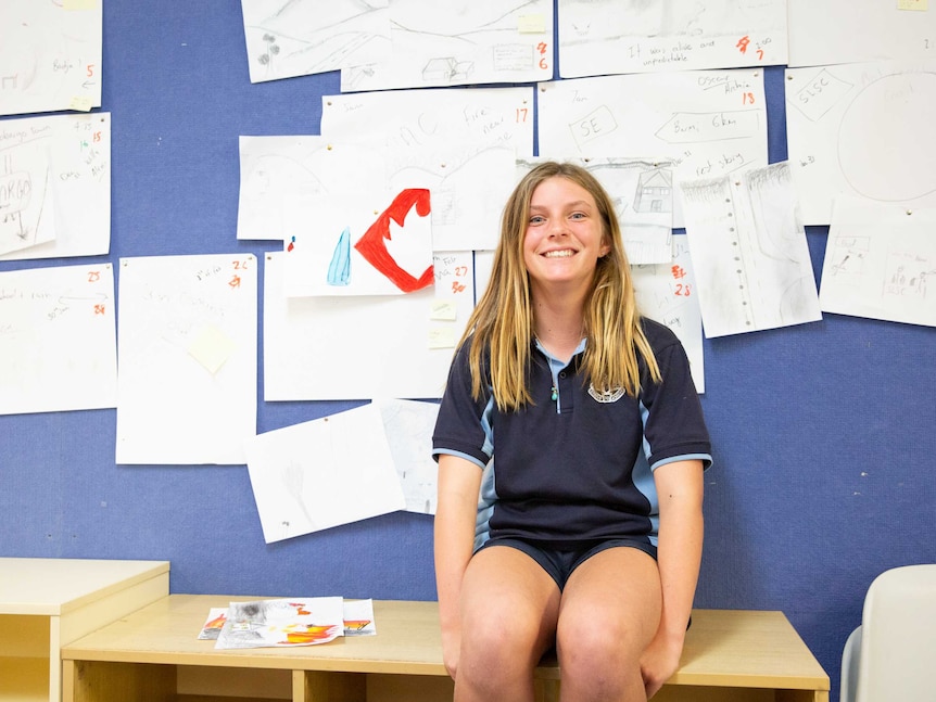 A girl in a school uniform sitting in front of a blue wall with children's art on it