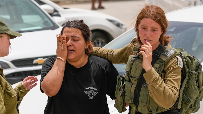 Two young women, one in a military uniform and the other in a military T-shirt, comfort each other as one cries.