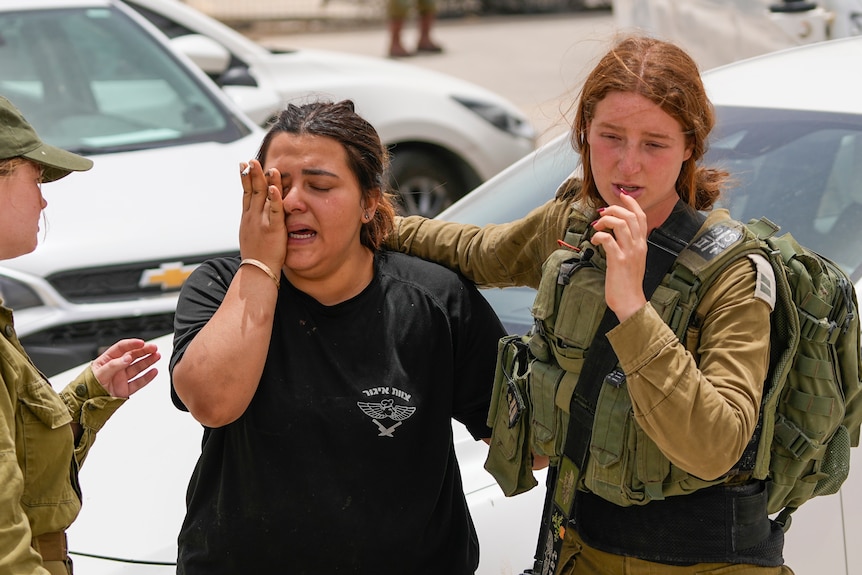 Two young women, one in a military uniform and the other in a military T-shirt, comfort each other as one cries.