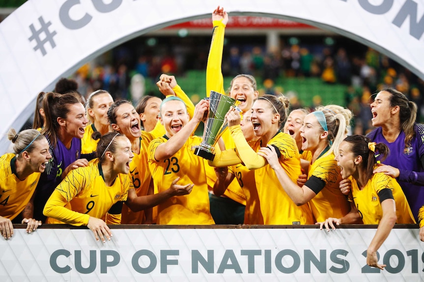 The Australian women's football team hold a trophy over their heads with glee.