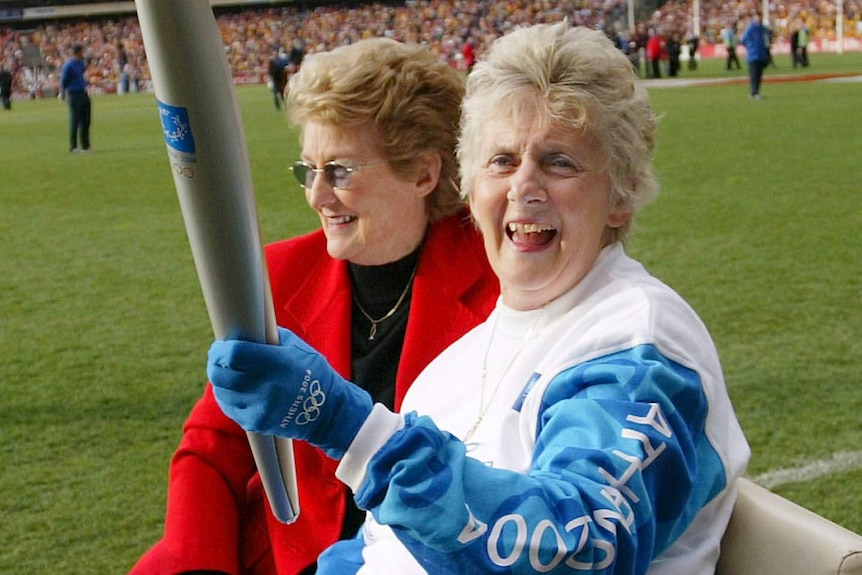Betty Cuthbert, seated, carries the Athens 2004 Olympic Torch with the Melbourne Cricket Ground in the background