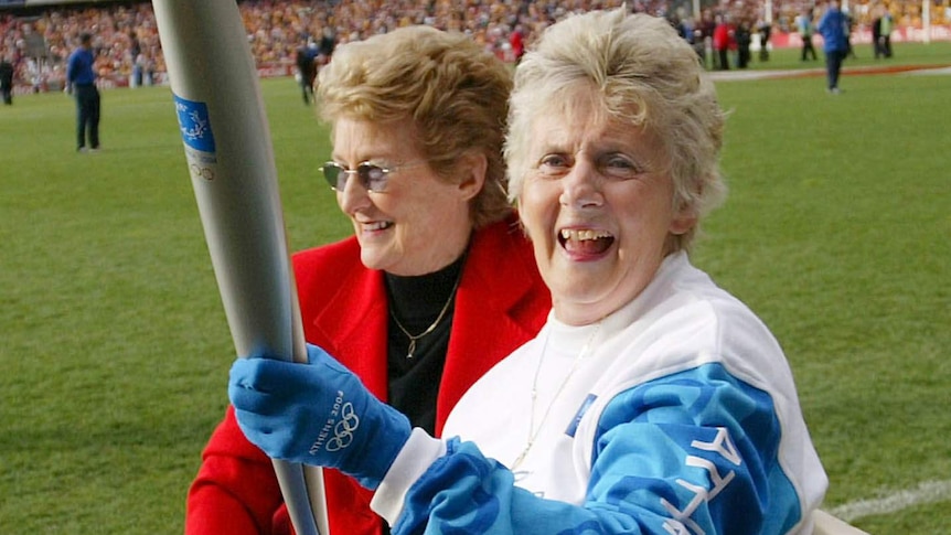 Betty Cuthbert, seated, carries the Athens 2004 Olympic Torch with the Melbourne Cricket Ground in the background