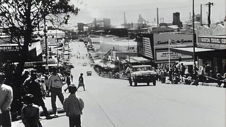 Wide black and white shot of a busy street looking downhill towards an industrial area.