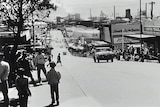 Wide black and white shot of a busy street looking downhill towards an industrial area.