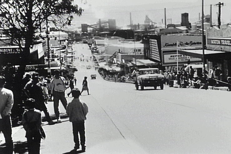 Wide black and white shot of a busy street looking downhill towards an industrial area.