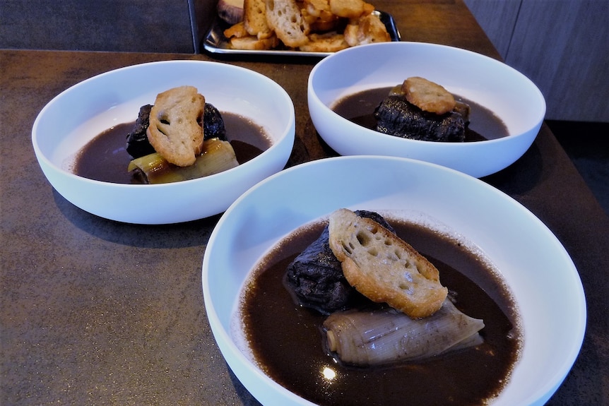 Three bowls with a dark looking sauce, bread, a green vegetable and a chunk of fish sit on a table.