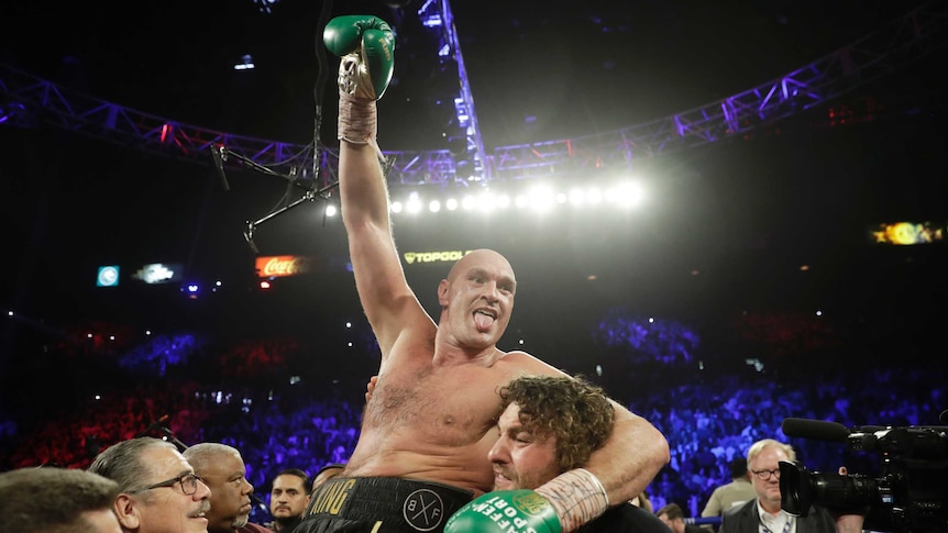 Tyson Fury raises a gloved fist and sticks his tongue out while his team lifts him on their shoulders after a heavyweight fight.