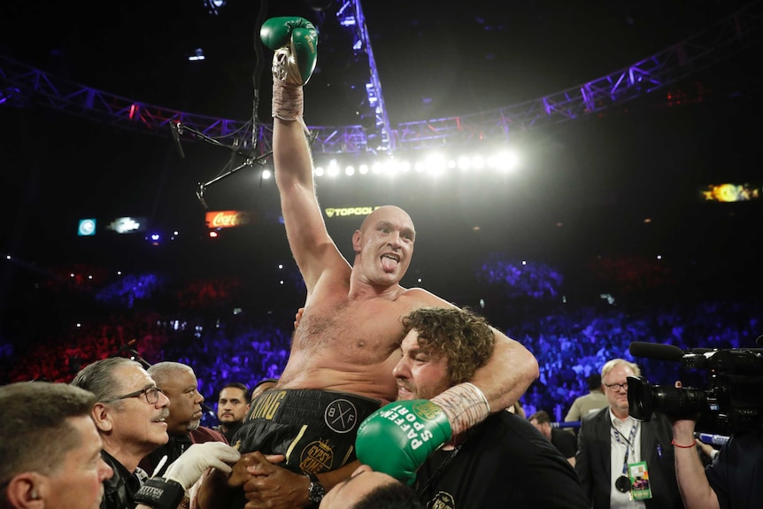 Tyson Fury raises a gloved fist and sticks his tongue out while his team lifts him on their shoulders after a heavyweight fight.