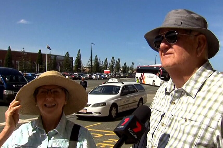 A woman with a large sun hat and a man wearing a fishing hat are interviewed in a car park.