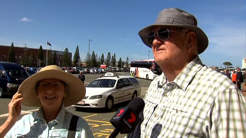 A woman with a large sun hat and a man wearing a fishing hat are interviewed in a car park.