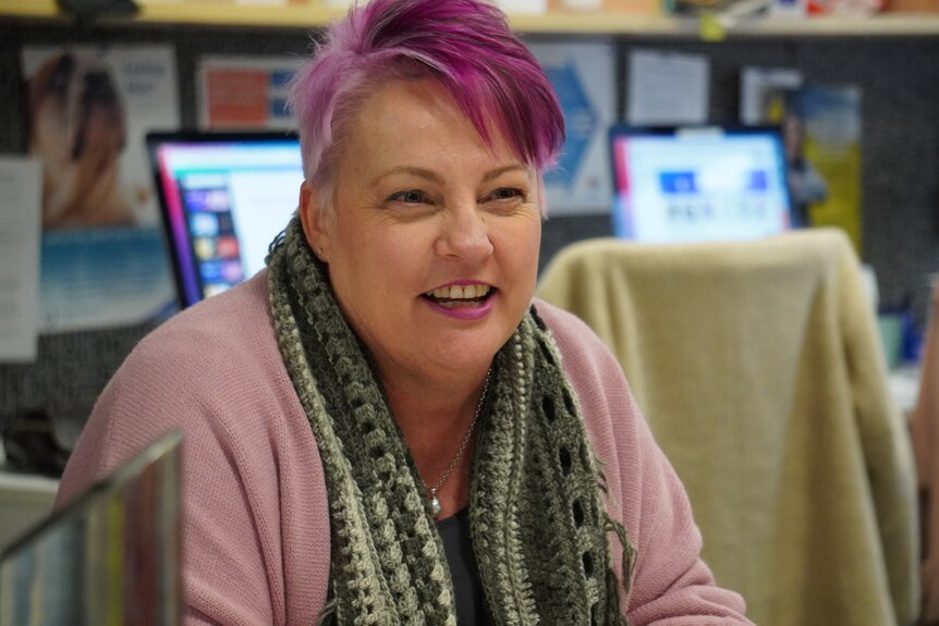 A head and shoulders shot of a woman with pink hair smiling while sitting down indoors.