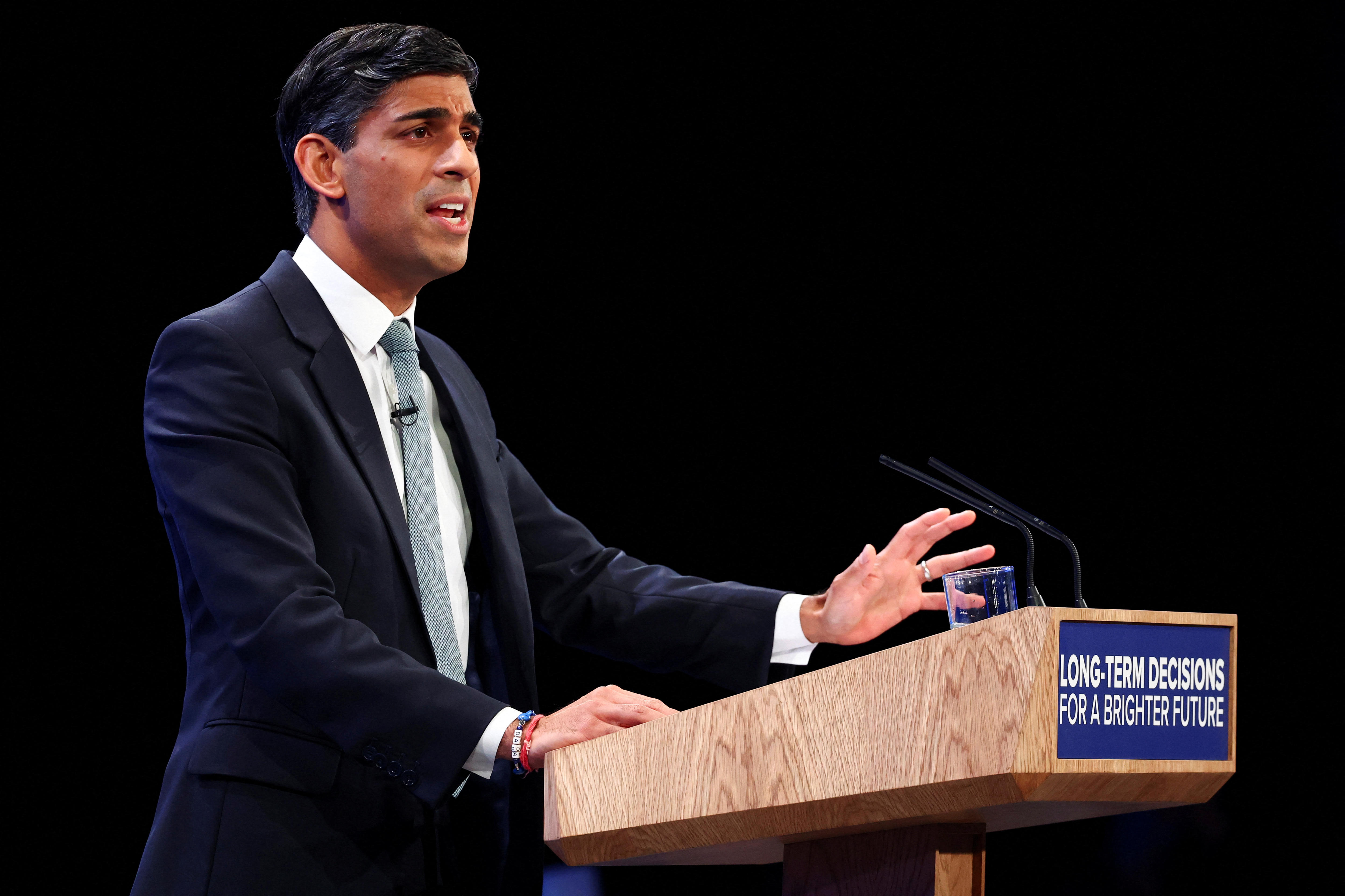 A confident-looking middle-aged man of Indian heritage in a suit speaks behind a woodern lectern.