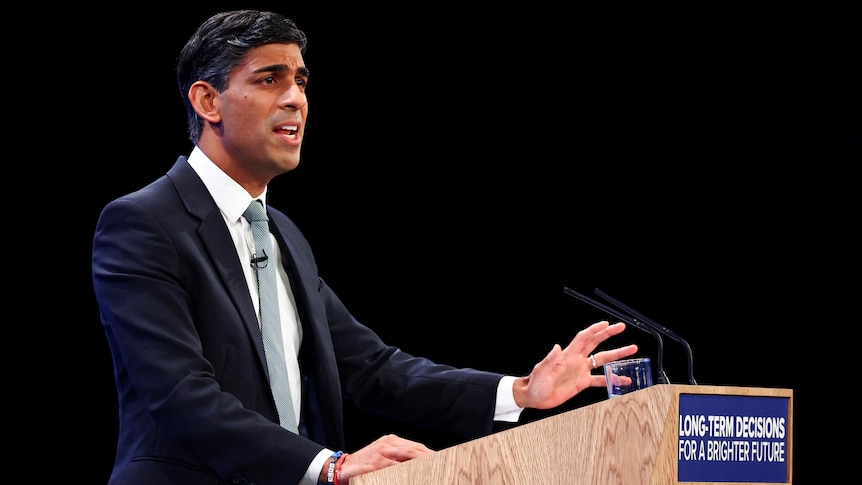 A confident-looking middle-aged man of Indian heritage in a suit speaks behind a woodern lectern.