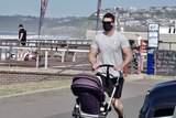 A man walking on a beachside footpath with a pram and wearing a black mask.