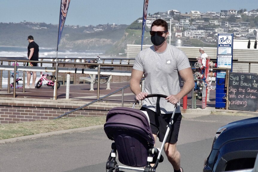 A man walking on a beachside footpath with a pram and wearing a black mask.