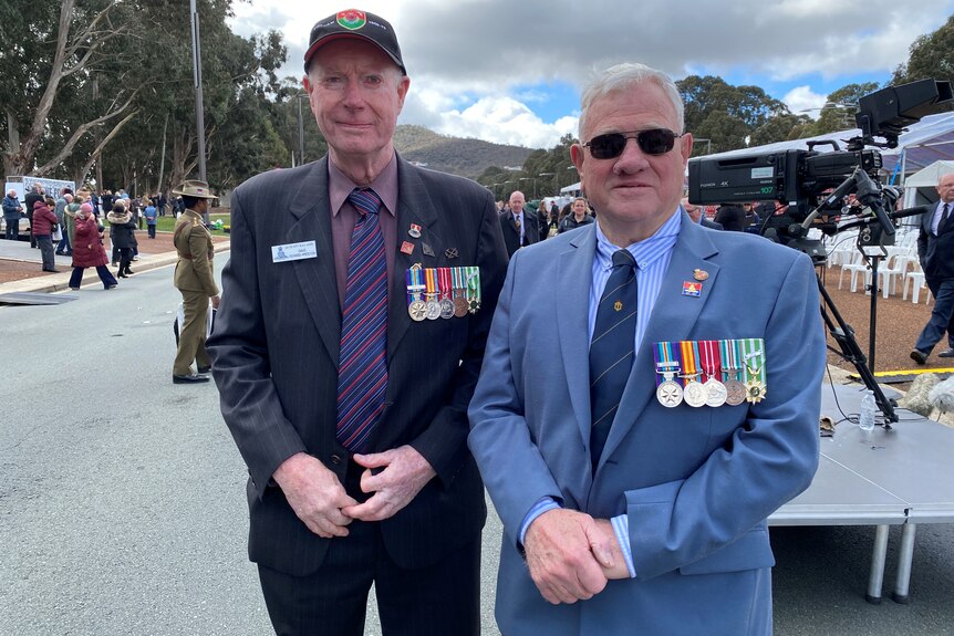 Dave and John stand wearing their medals, outside near the memorial.