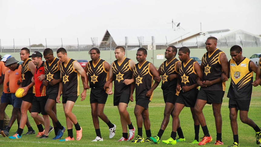 Line of men in football uniforms with linked arms walking on a green football pitch.