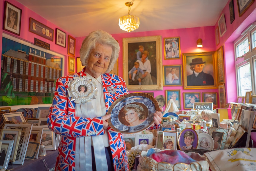 A woman in a Union Jack flag jacket holds a Princess Diana plate in a bright pink room.