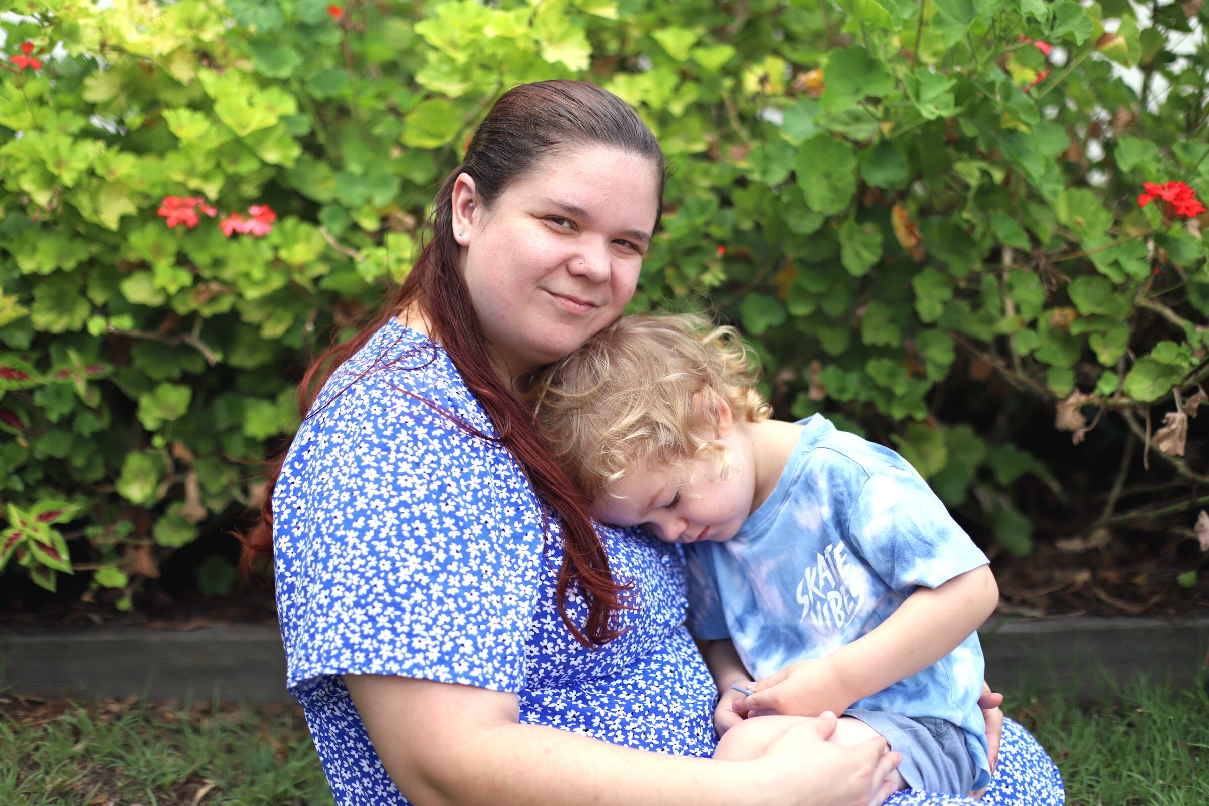 Tasha Torzsa smiles while holding her young son in her arms. A green shrub with pink flowers is behind them.