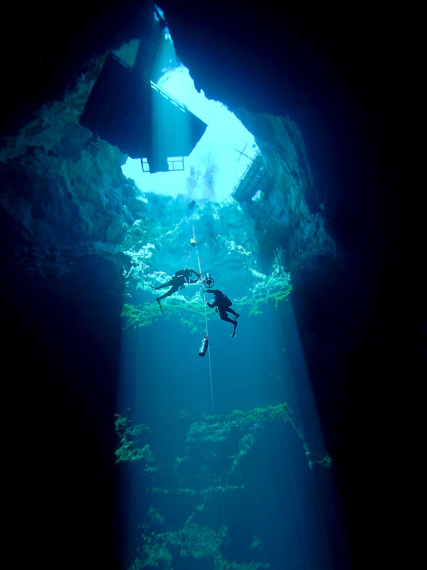 Two scuba divers traverse down a line in crystal clear water under a large light beam from above.