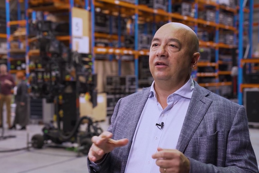 An image of a man in a suit gesticulating with film studio equipment in the background