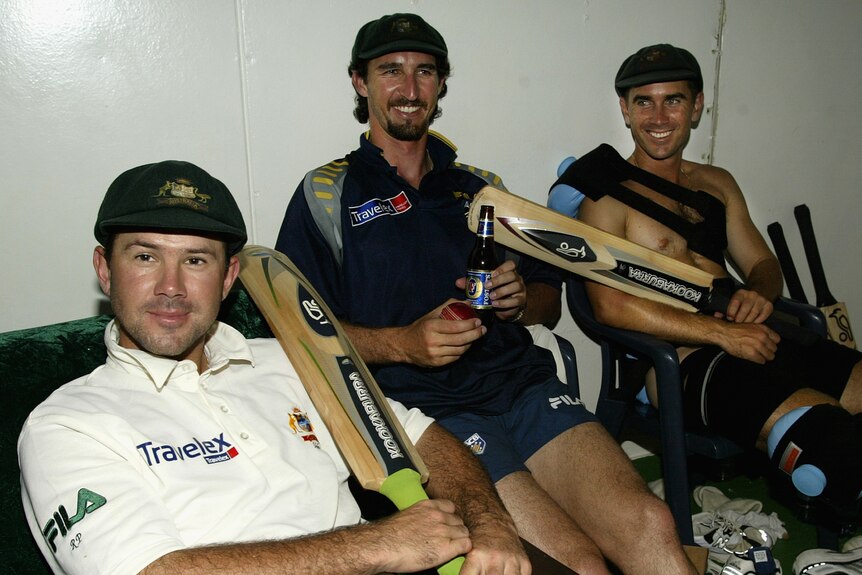 Three men sit with cricket gear in a changeroom