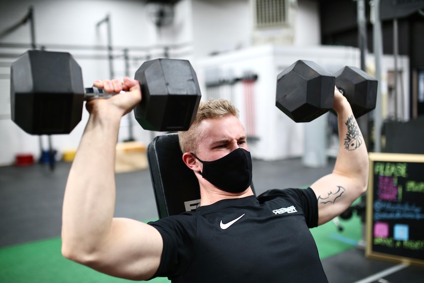 Brayden wearing a black t-shirt, sitting on a bench in a gym, lifting two weights - one with each hand - above head