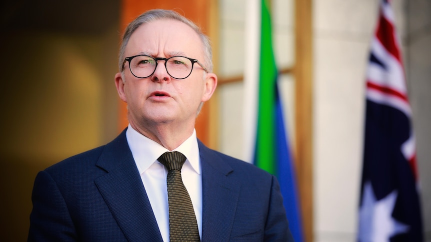 An earnest-looking man in a suit with grey hair and black glasses speaks in front of a pair of flags.