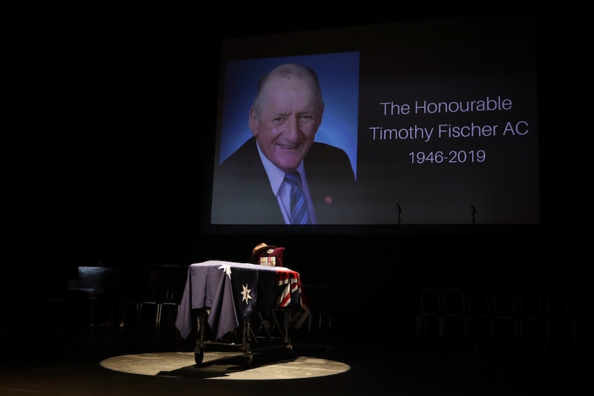 A coffin on a stage, draped in an Australian flag. An image of Tim Fischer 1946 - 2019 is on the big screen.