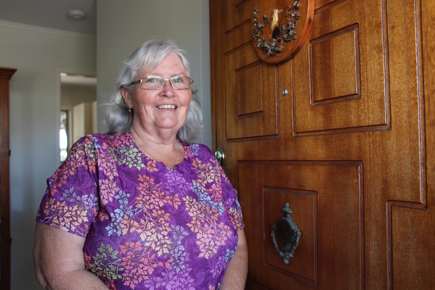 A smiling older woman with fair skin and white hair standing by her open front door.