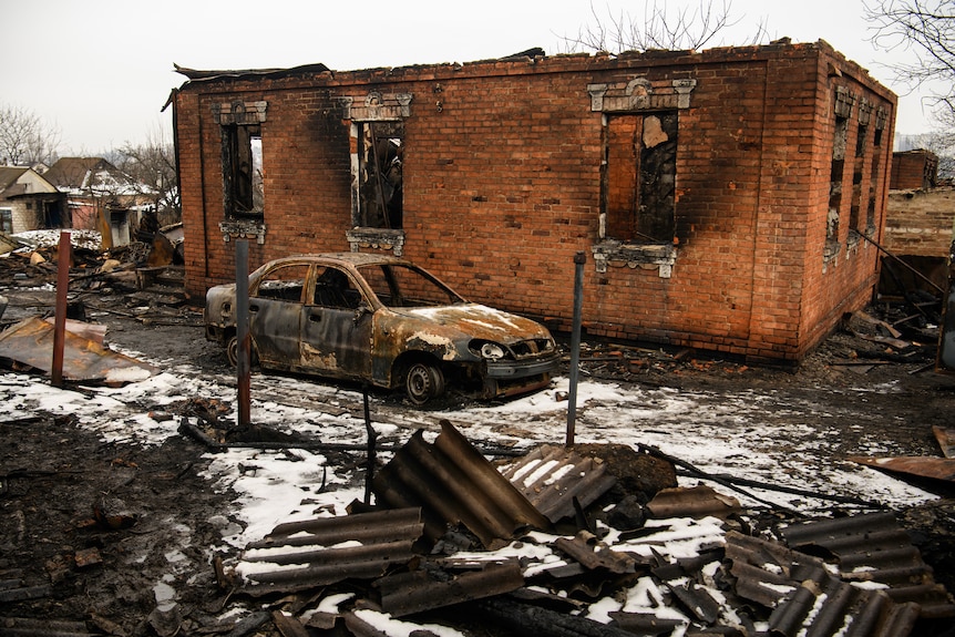 A burnt out car sits next to a burnt out building and there is rubble all around.