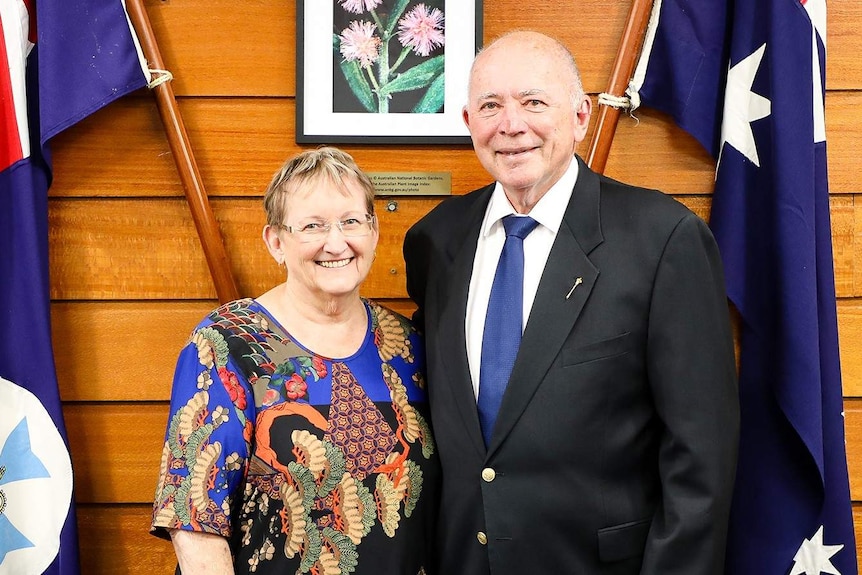Woman and bald man in suit and tie standing between Australian and Queensland flags