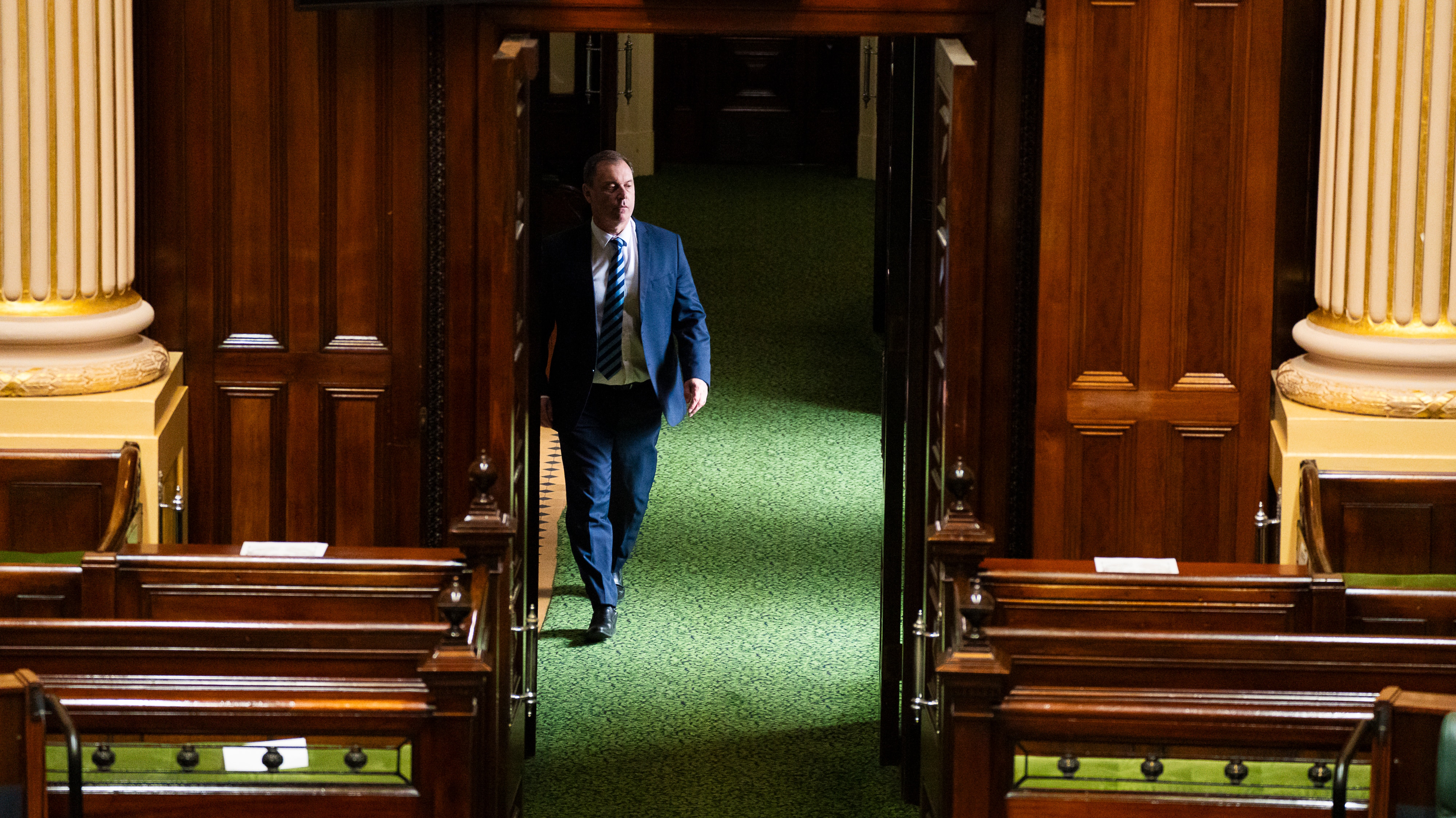A man in a blue suit, white shirt and blue striped tie walks over green carpet towards an open set of wooden doors.
