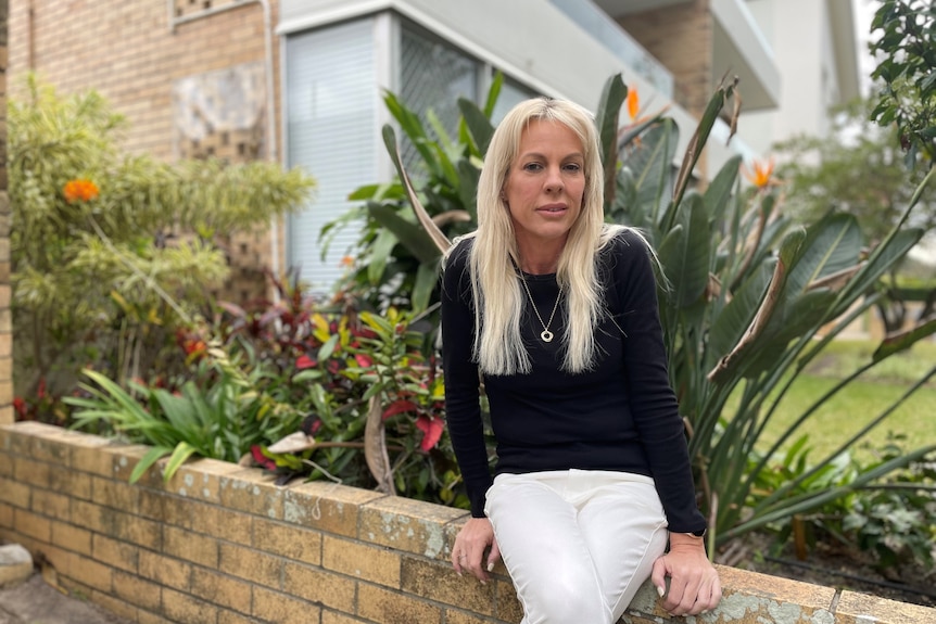 Woman with long blonde hair wearing a black top and white pants sitting on a low brick wall with plants behind her.