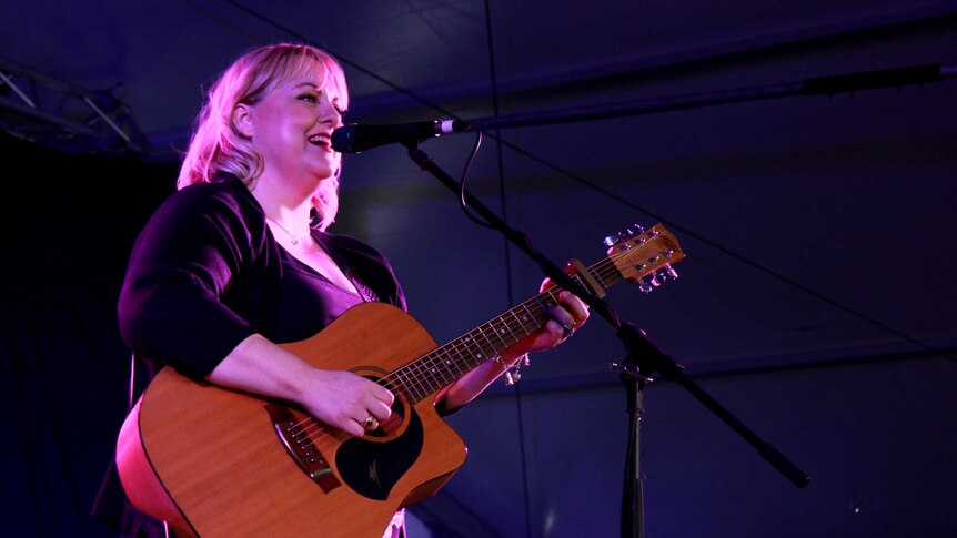 A female singer holding an acoustic guitar on stage shot from below in a purple light