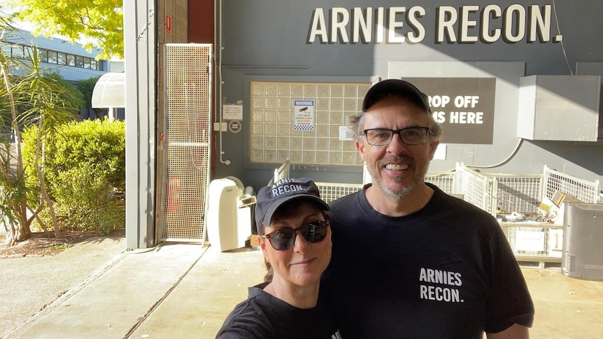 A woman wearing glasses stands next to a man in a black t-shirt in a warehouse. 