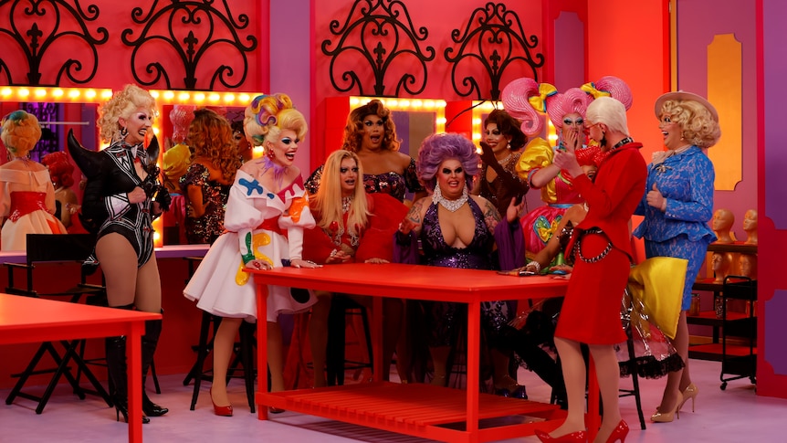 A group of drag queens huddled together around a table, the cast of RuPaul's Drag Race Down Under in episode 1