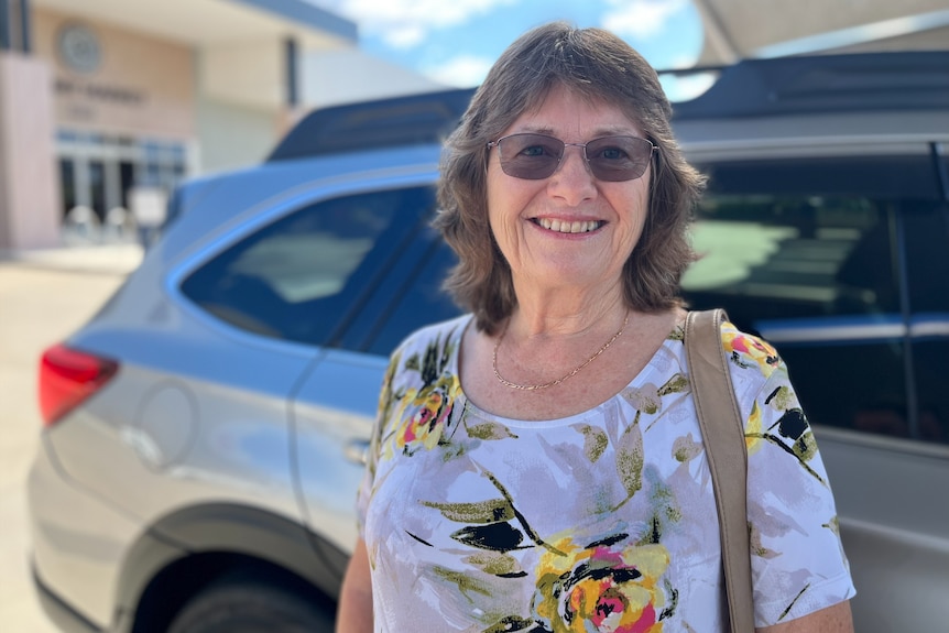 A smiling older woman stands in front of a car.