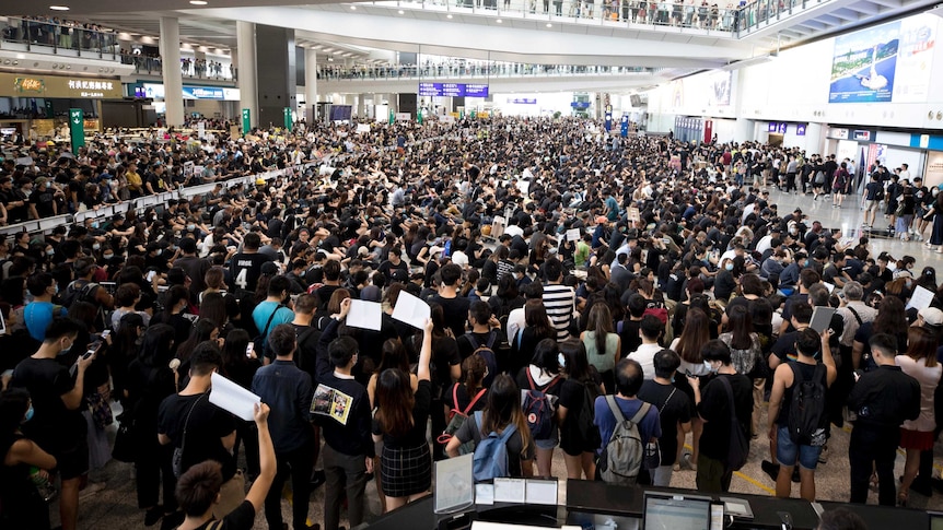 A large crowd of people stand inside an airport some are holding up signs