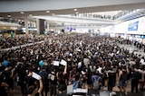 A large crowd of people stand inside an airport some are holding up signs