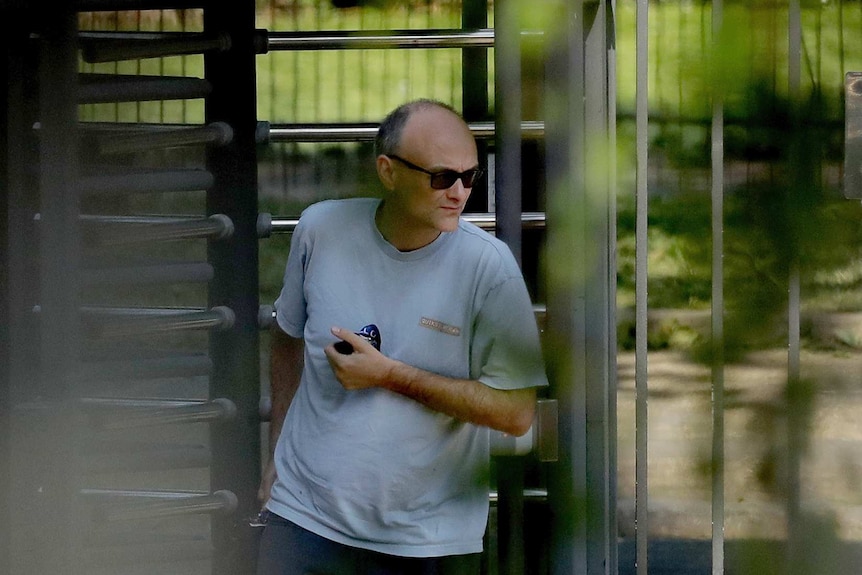 A balding middle-aged man wearing a grey T-shirt walks through a revolving gate in a wooded area.