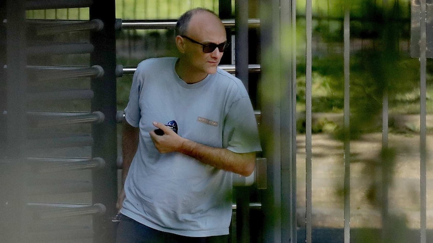 A balding middle-aged man wearing a grey T-shirt walks through a revolving gate in a wooded area.