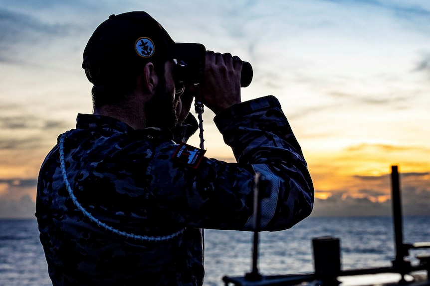 Oficial naval en un barco mirando a través de binoculares al atardecer. 