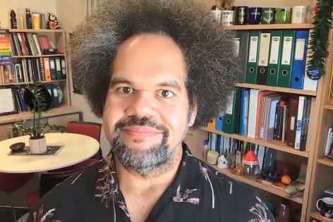 A fijian man with a large afro wearing a colourful t-shirt in his office surrounded by books