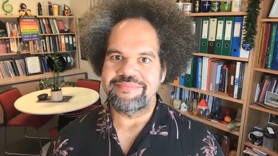 A fijian man with a large afro wearing a colourful t-shirt in his office surrounded by books