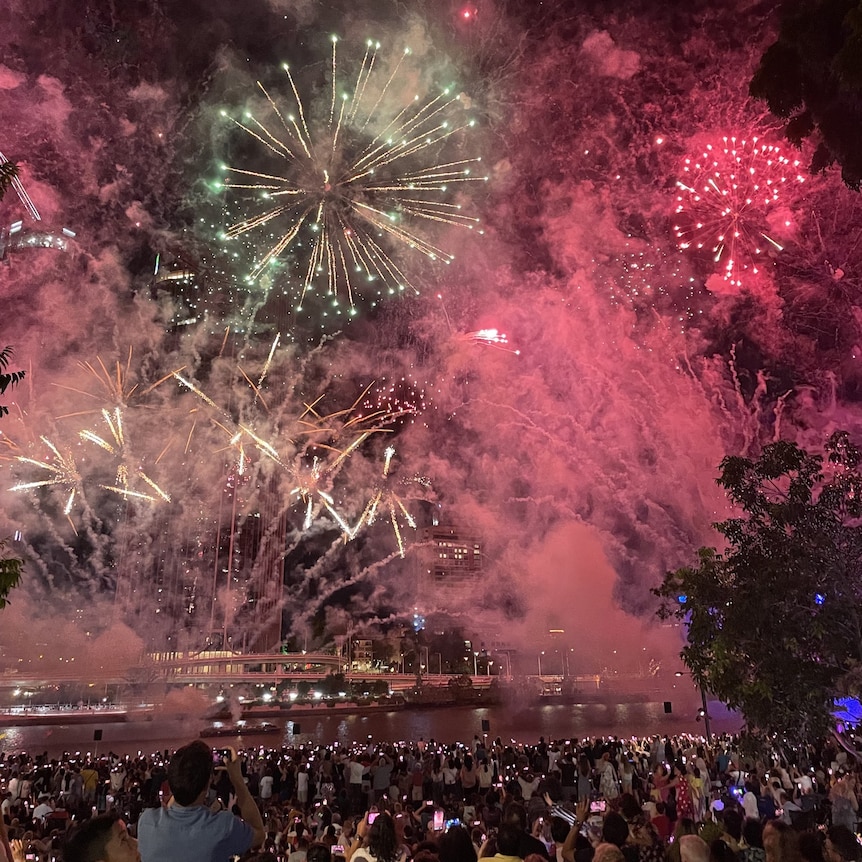 A wide image of a crowd of people looking up at fireworks in the night sky above the Brisbane River.