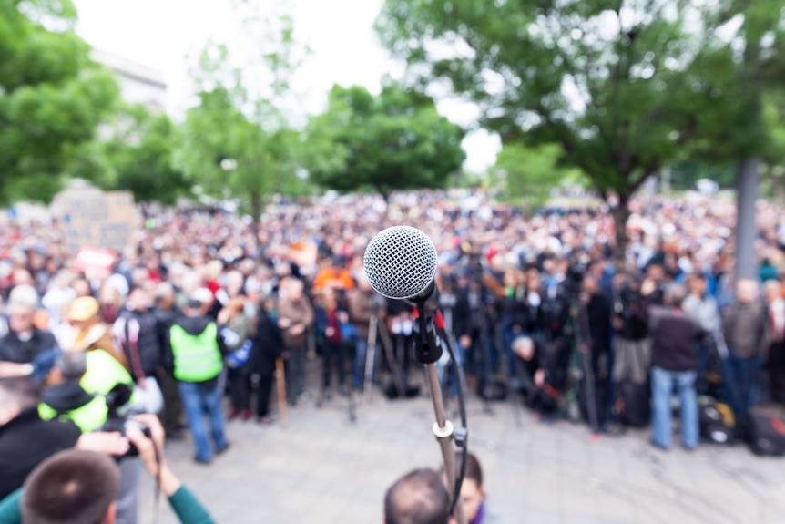 At centre of image is a microphone. Behind it a large, blurred crowd of people. Trees surround them.