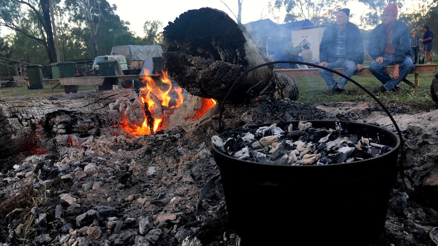In the foreground a cast iron camp oven sits in a fire with coals on top while two men watch on in the background