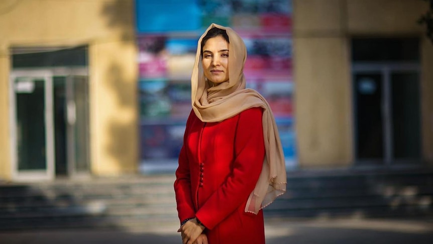 A young Afghan woman in a red long sleeved dress and cream head scarf looking strong 